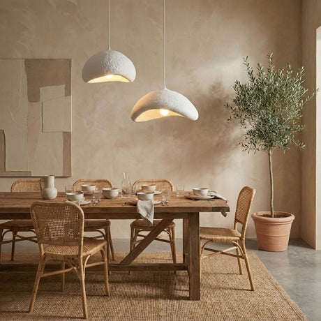 Een sfeervolle, neutrale eetkamer met een robuuste houten tafel, rotan stoelen, witte organische hanglampen en een olijfboom.